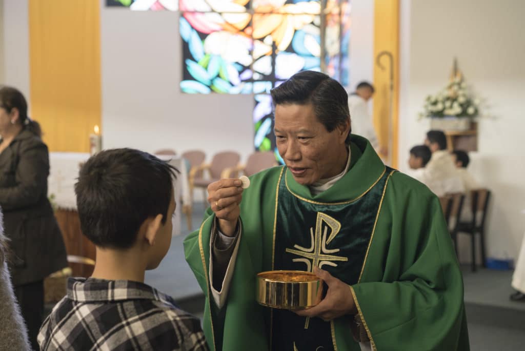 Fr Remy Lam Son Bui, parish priest of the nearby parish of All Saints, Liverpool, holds up the host as he gives Communion during Mass at St Francis Xavier, Lurnea. Photo: Patrick J Lee